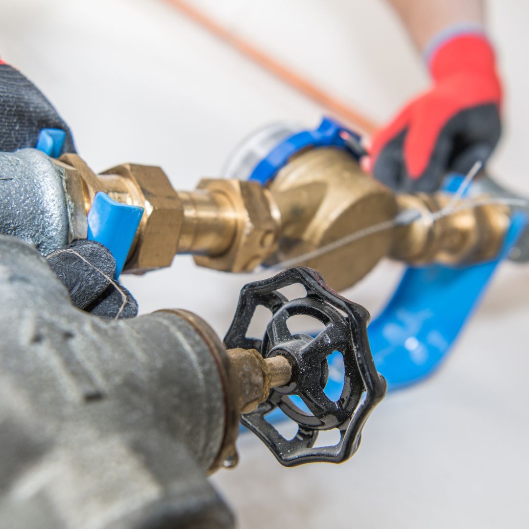 Detalhe de instalação de cavalete de água com registro de latão e hidrômetro. Técnico com luvas de proteção realizando manutenção na rede hidráulica.